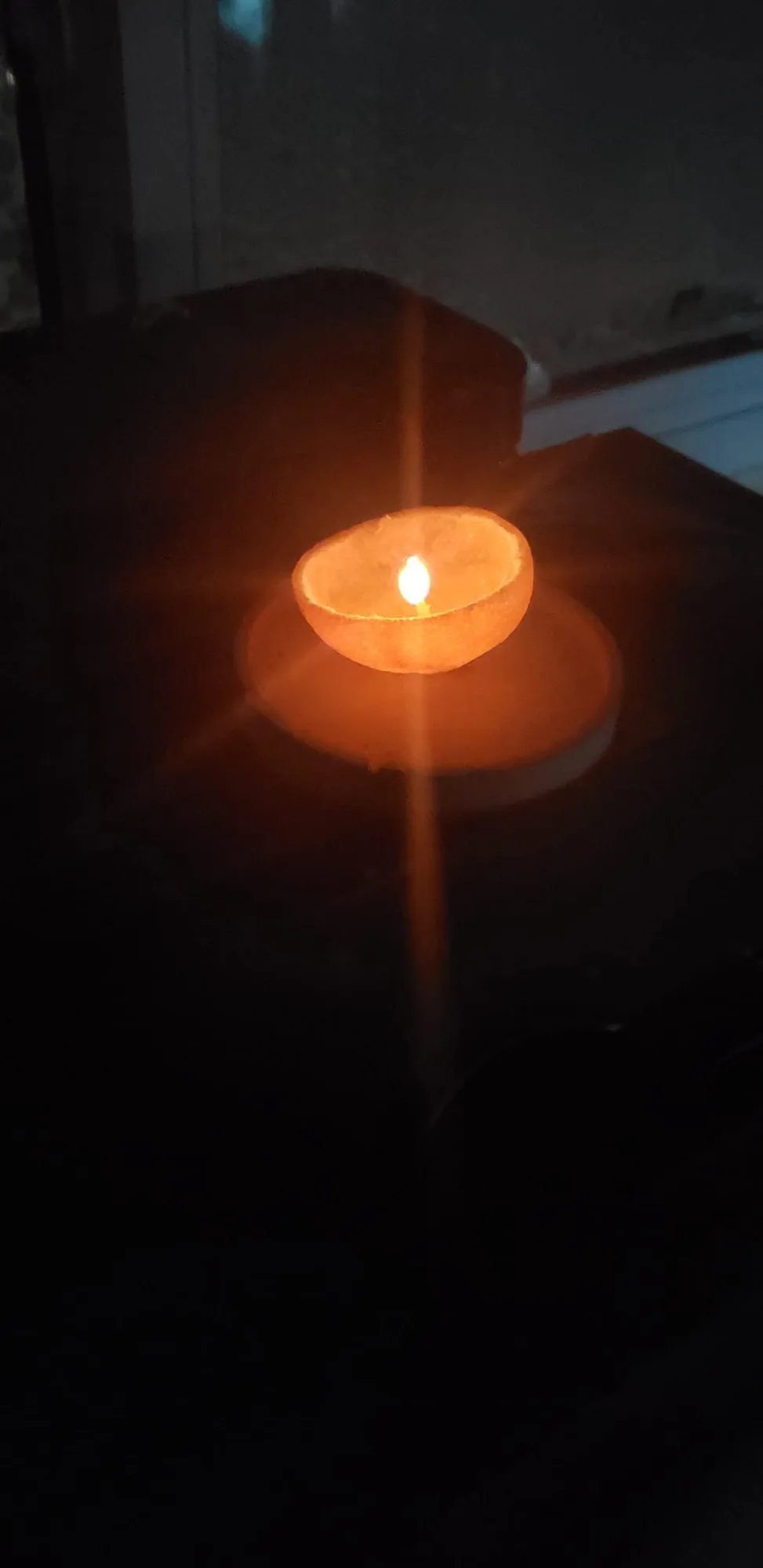 A glowing orange peel candle burning in a dark room, casting a warm amber light with its reflection visible on a windowpane.